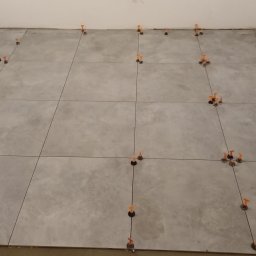 Piotr Korbel Vertikor - Szare płytki podłogowe w trakcie układania, z pomarańczowymi i niebieskimi klinami poziomującymi w spoinach, widok z góry na częściowo wykończoną powierzchnię.