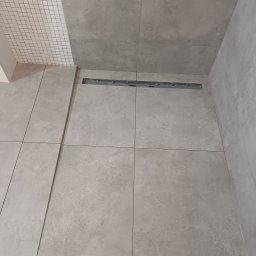 Piotr Korbel Vertikor - Ułożone szare płytki podłogowe w kabinie prysznicowej z odpływem liniowym i fragmentem ściany wyłożonej mozaiką.