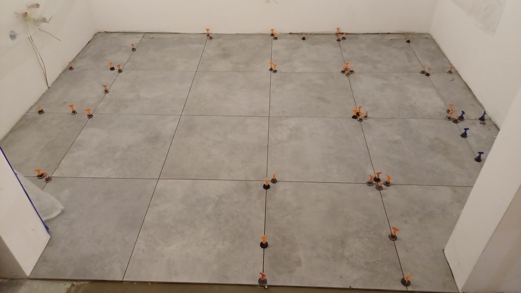 Szare płytki podłogowe w trakcie układania, z pomarańczowymi i niebieskimi klinami poziomującymi w spoinach, widok z góry na częściowo wykończoną powierzchnię.