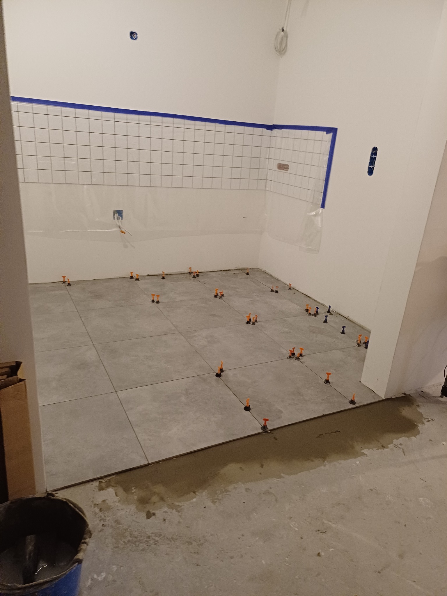 Układanie szarych płytek podłogowych w łazience z użyciem klipsów poziomujących, widoczna ściana z białymi płytkami zabezpieczona niebieską taśmą malarską.