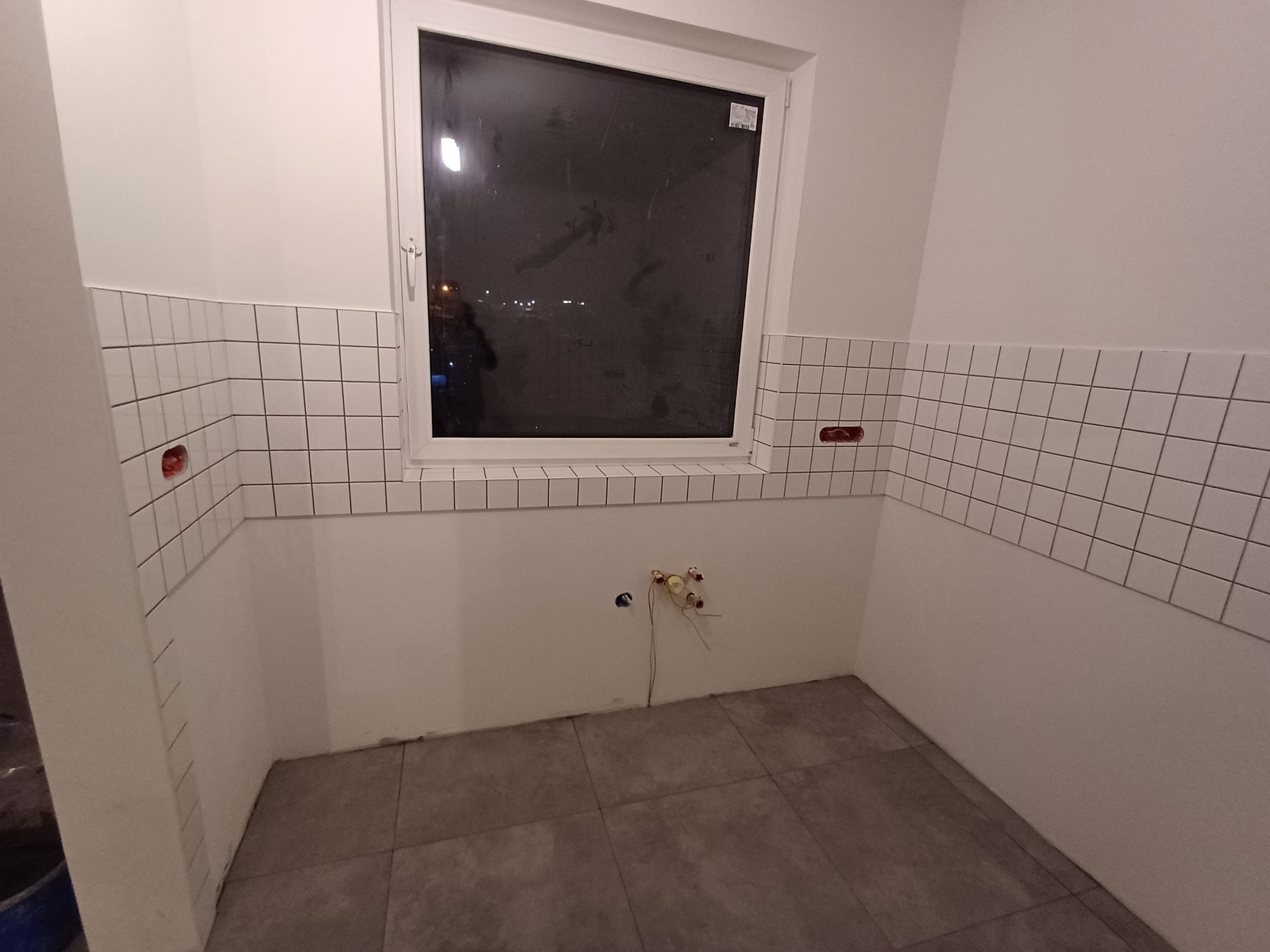 Wnętrze łazienki w trakcie prac wykończeniowych, widoczne ułożone białe płytki na ścianach do pewnej wysokości, szare płytki podłogowe, okno z widokiem na nocne miasto oraz wystające instalacje...