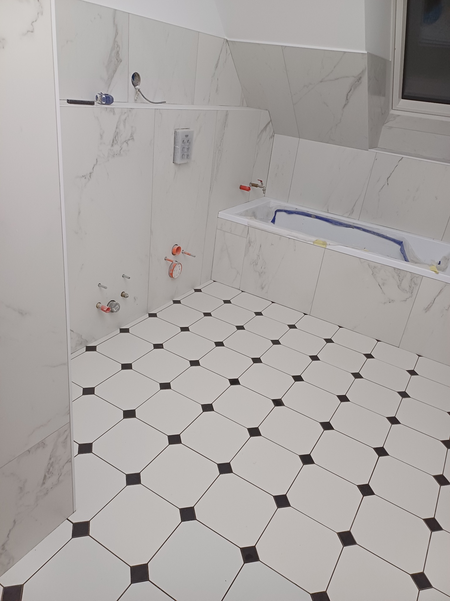Łazienka w trakcie wykańczania: białe płytki imitujące marmur na ścianach, czarno-biała podłoga w karo, przygotowana wanna zabezpieczona folią, widoczne instalacje wodne i elektryczne.