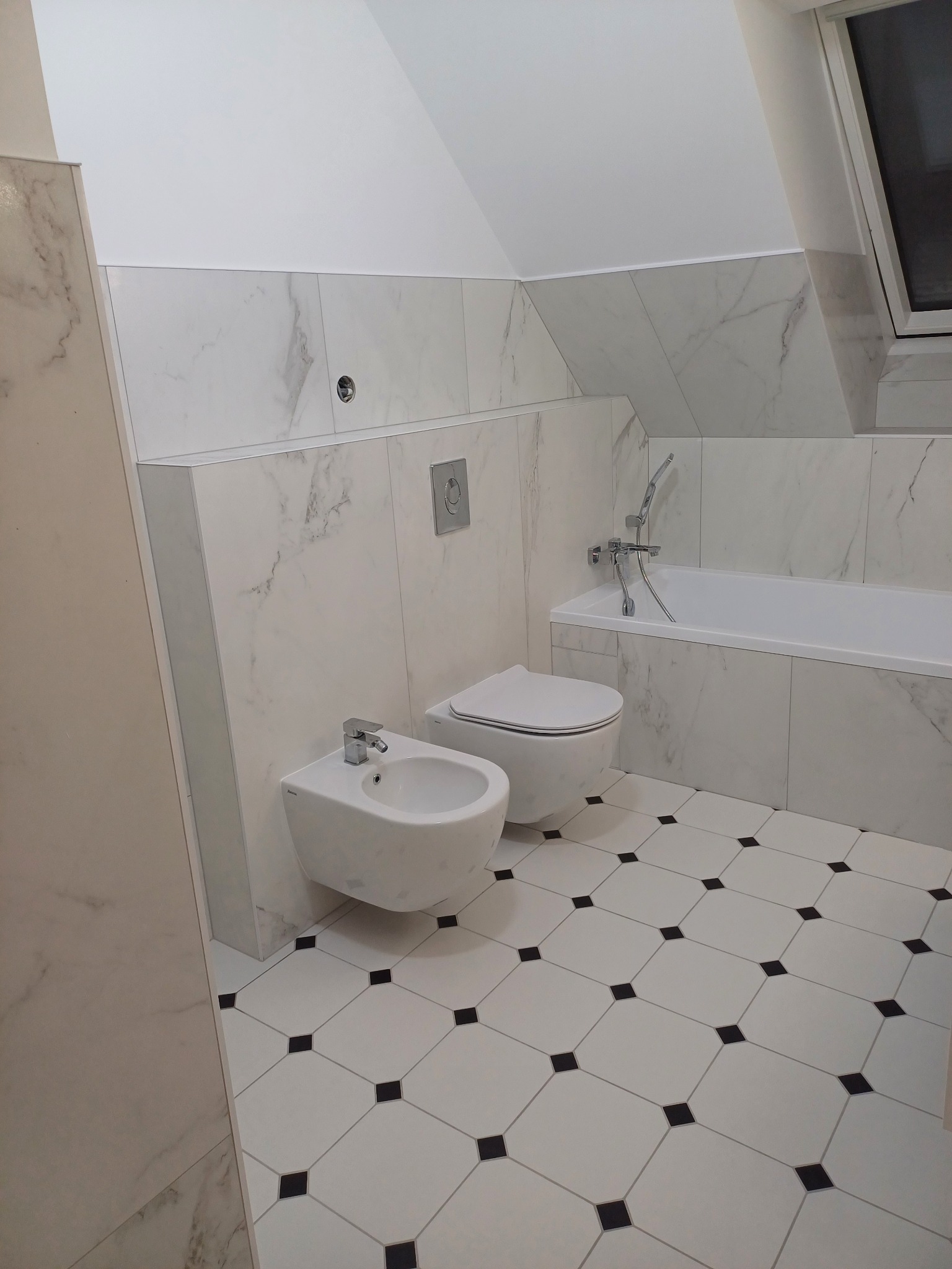 Nowoczesna łazienka na poddaszu z biało-czarną podłogą w karo, białe płytki imitujące marmur na ścianach, wisząca toaleta i bidet oraz wanna z chromowaną armaturą.