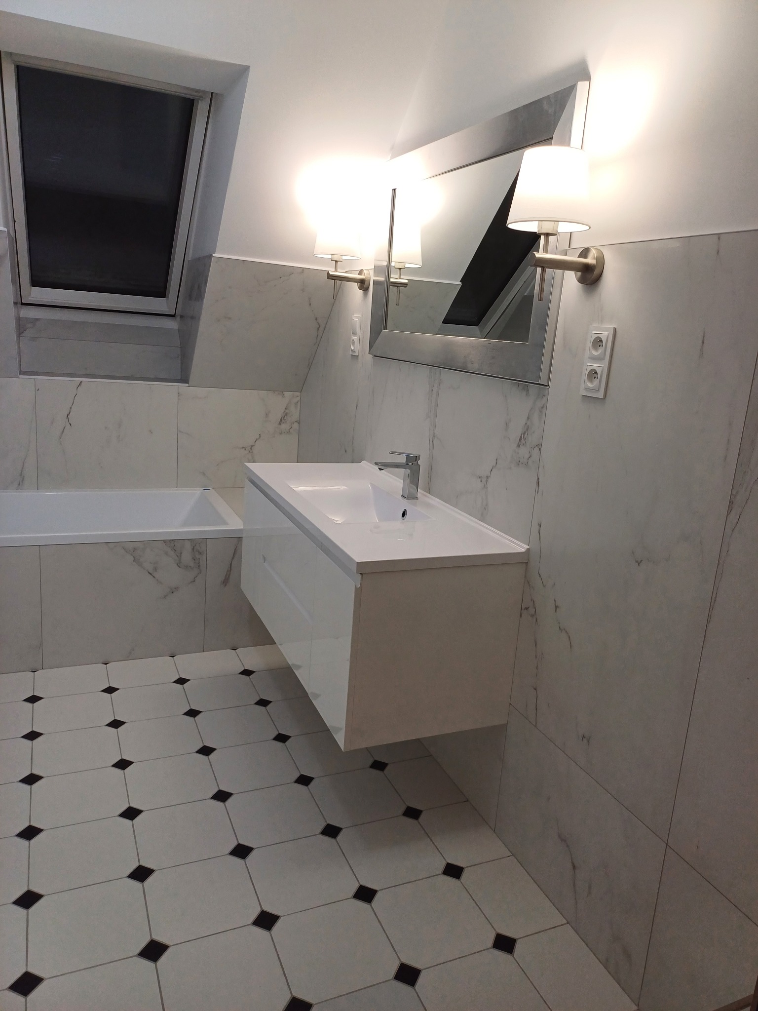 Nowoczesna łazienka z biało-szarymi marmurowymi płytkami, białą wiszącą szafką z umywalką, lustrem z oświetleniem i podłogą w biało-czarną szachownicę, widok na okno dachowe.