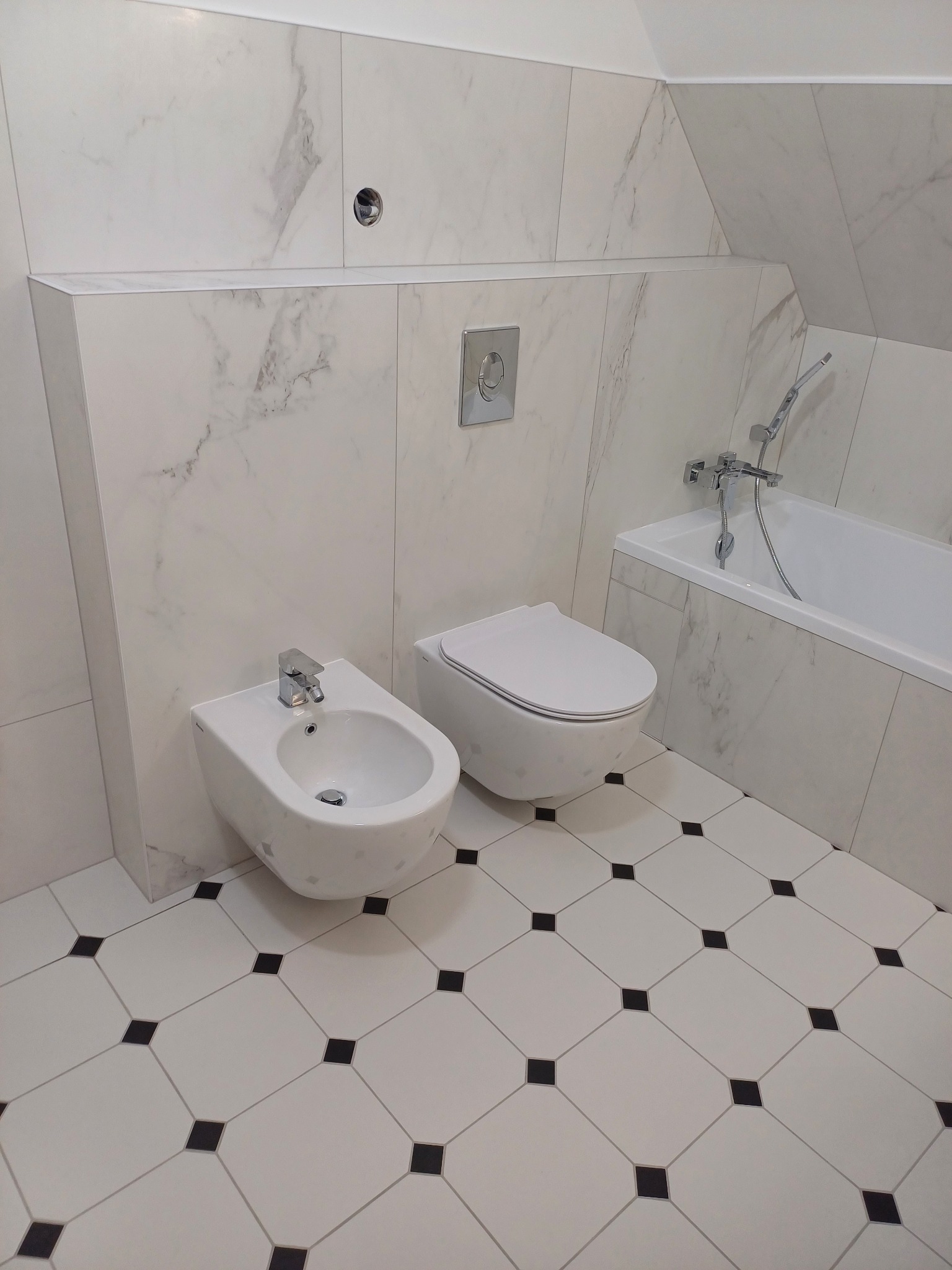 Nowoczesna łazienka z marmurowymi płytkami, białą armaturą (bidet i toaleta) oraz podłogą w biało-czarną szachownicę, widok na baterię wannową.