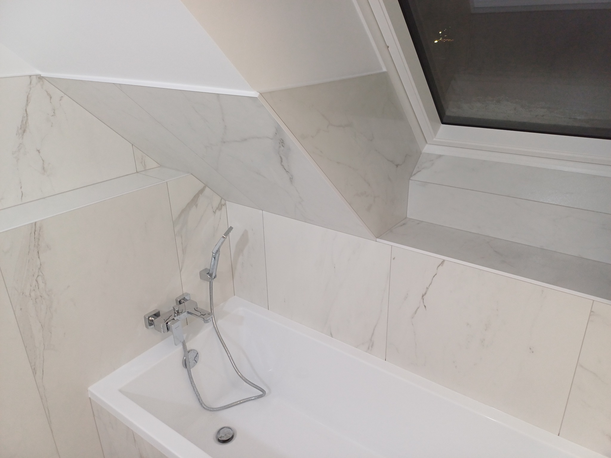 Nowoczesna łazienka na poddaszu z białą wanną, chromowaną baterią i kabiną prysznicową oraz marmurowymi płytkami ułożonymi pod skosem, oświetlona przez okno dachowe.