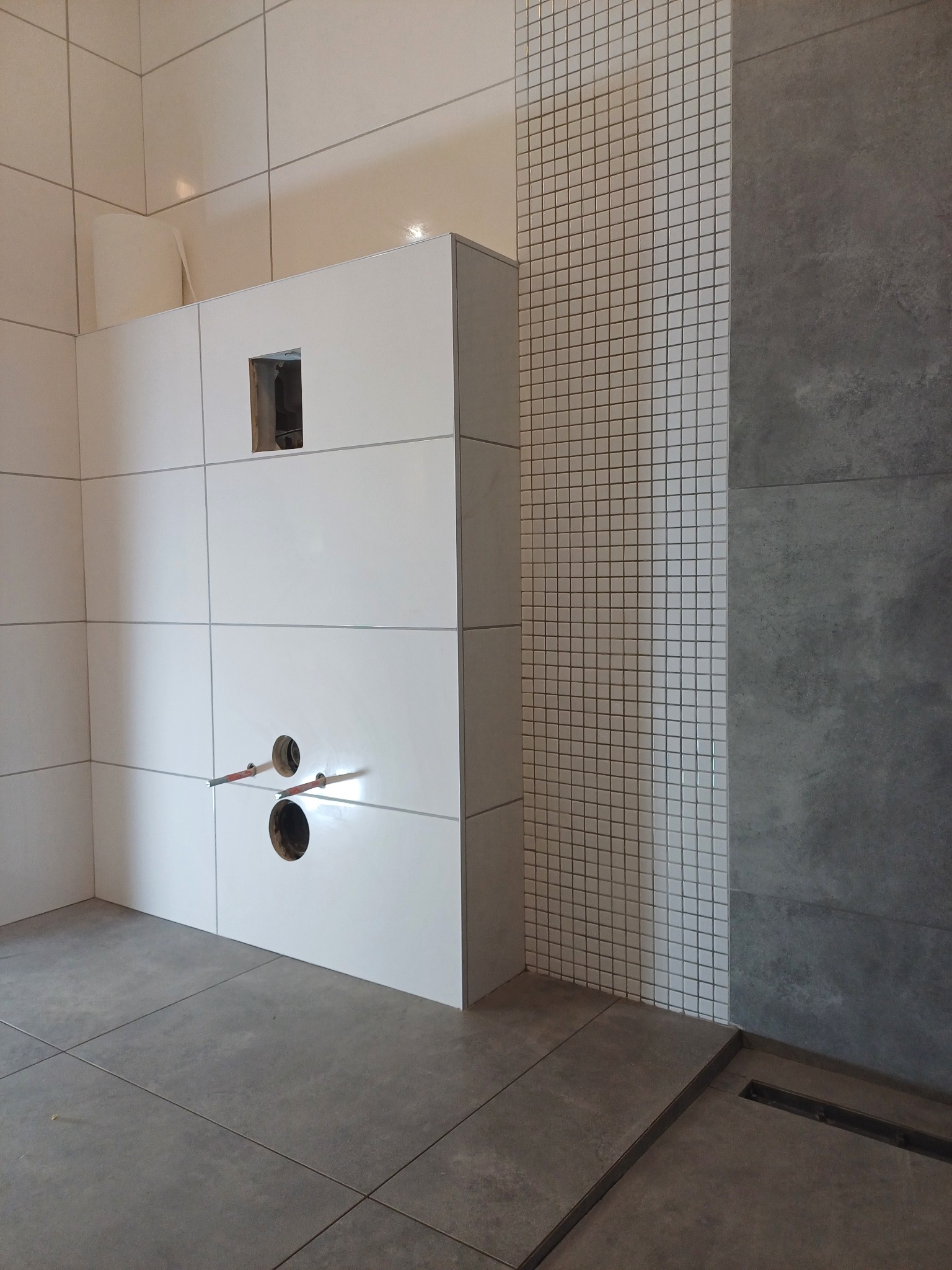 Wnętrze łazienki w trakcie prac wykończeniowych; widoczne białe, prostokątne płytki na ścianach, fragment ściany z mozaiki oraz szare płytki podłogowe z odpływem liniowym; instalacja wodna wystaje...