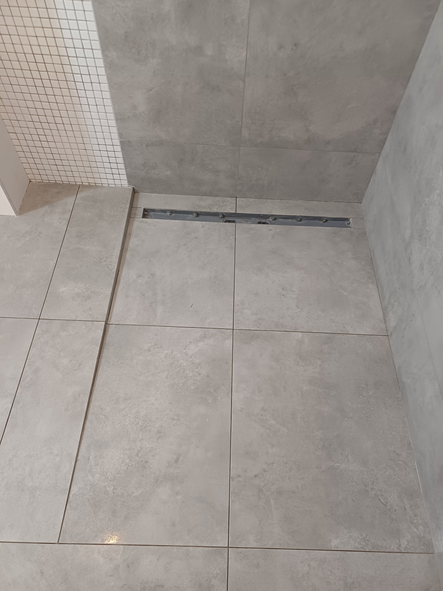 Ułożone szare płytki podłogowe w kabinie prysznicowej z odpływem liniowym i fragmentem ściany wyłożonej mozaiką.