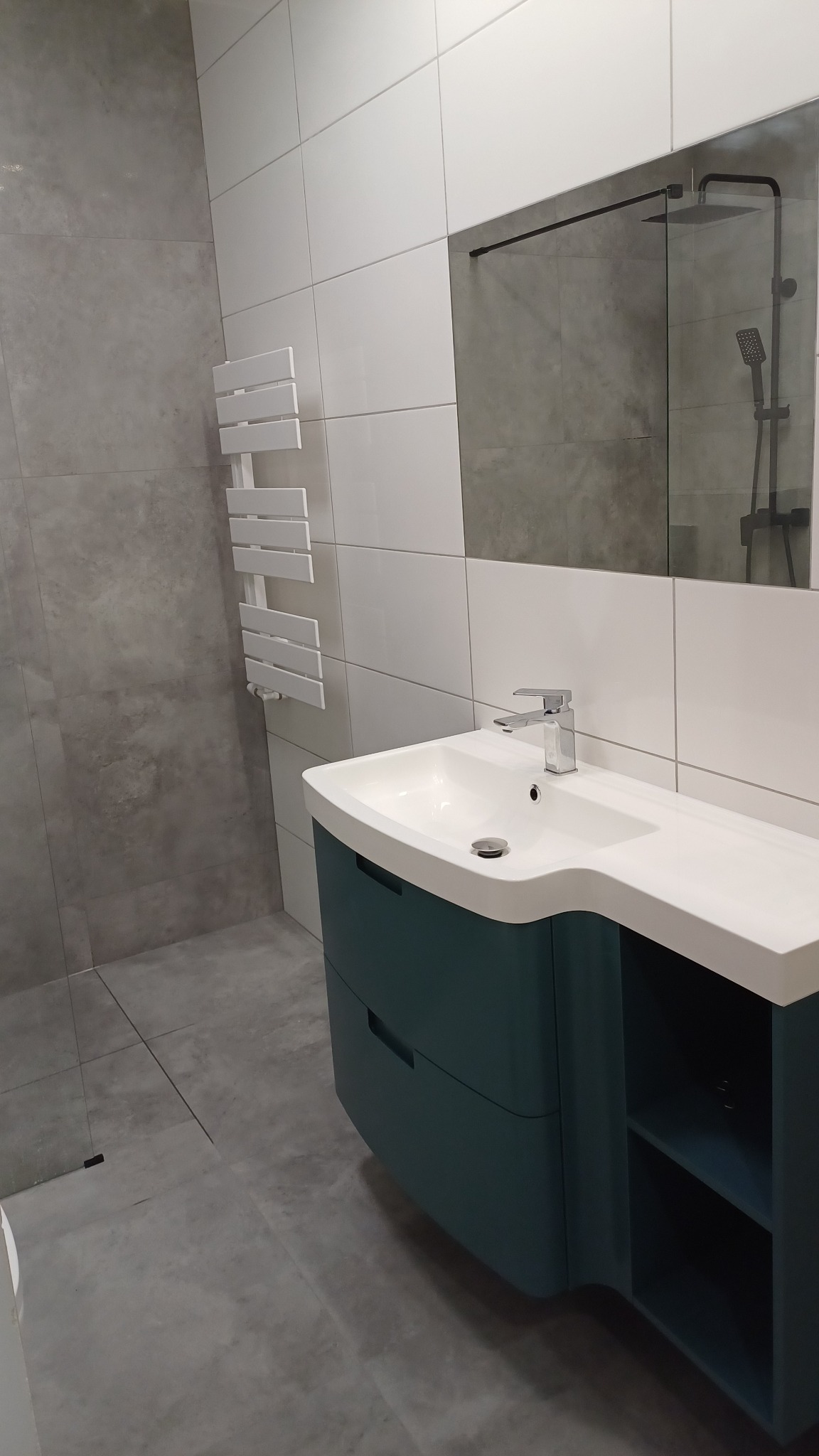 Nowoczesna łazienka z umywalką na szafce w odcieniach turkusu, białe i szare płytki na ścianach, grzejnik drabinkowy i lustro z widoczną kabiną prysznicową.