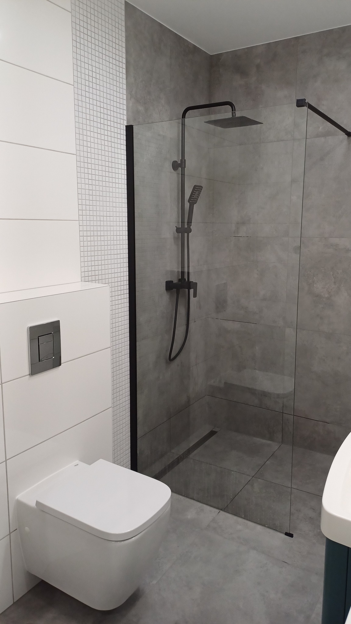 Nowoczesna łazienka z szarą ścianą imitującą beton, białą mozaiką i podwieszaną toaletą, kabina prysznicowa typu walk-in z czarną armaturą.