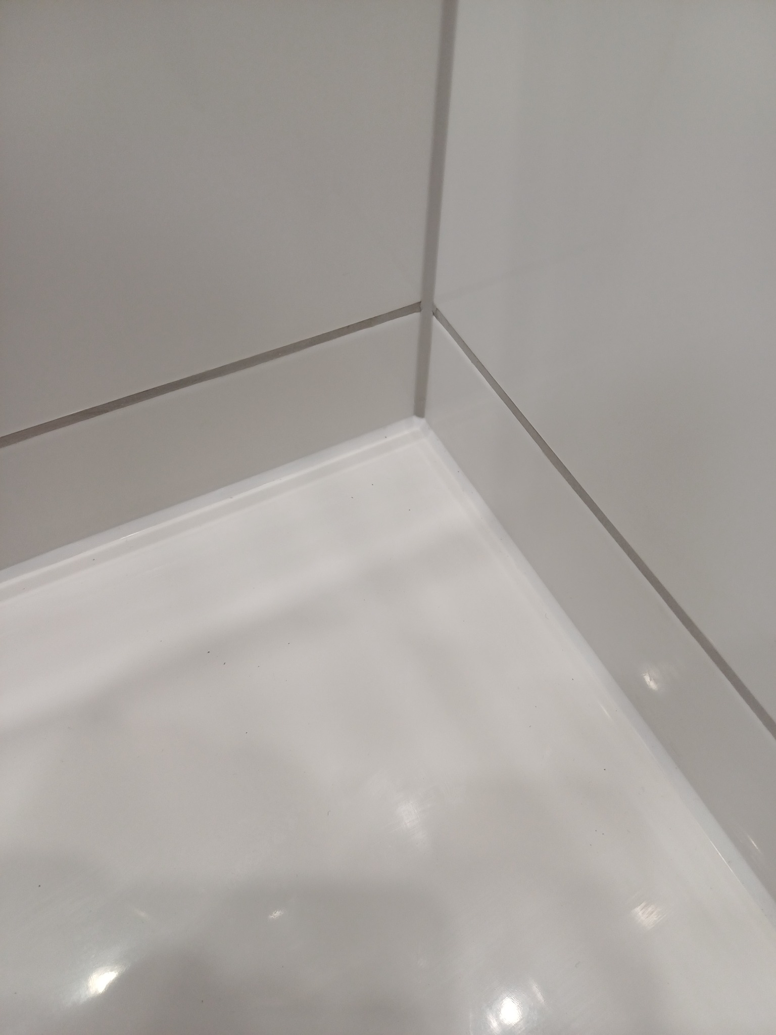 Narożnik kabiny prysznicowej wykończony białymi, błyszczącymi płytkami z delikatnymi fugami, widoczne połączenie ściany i brodzika.