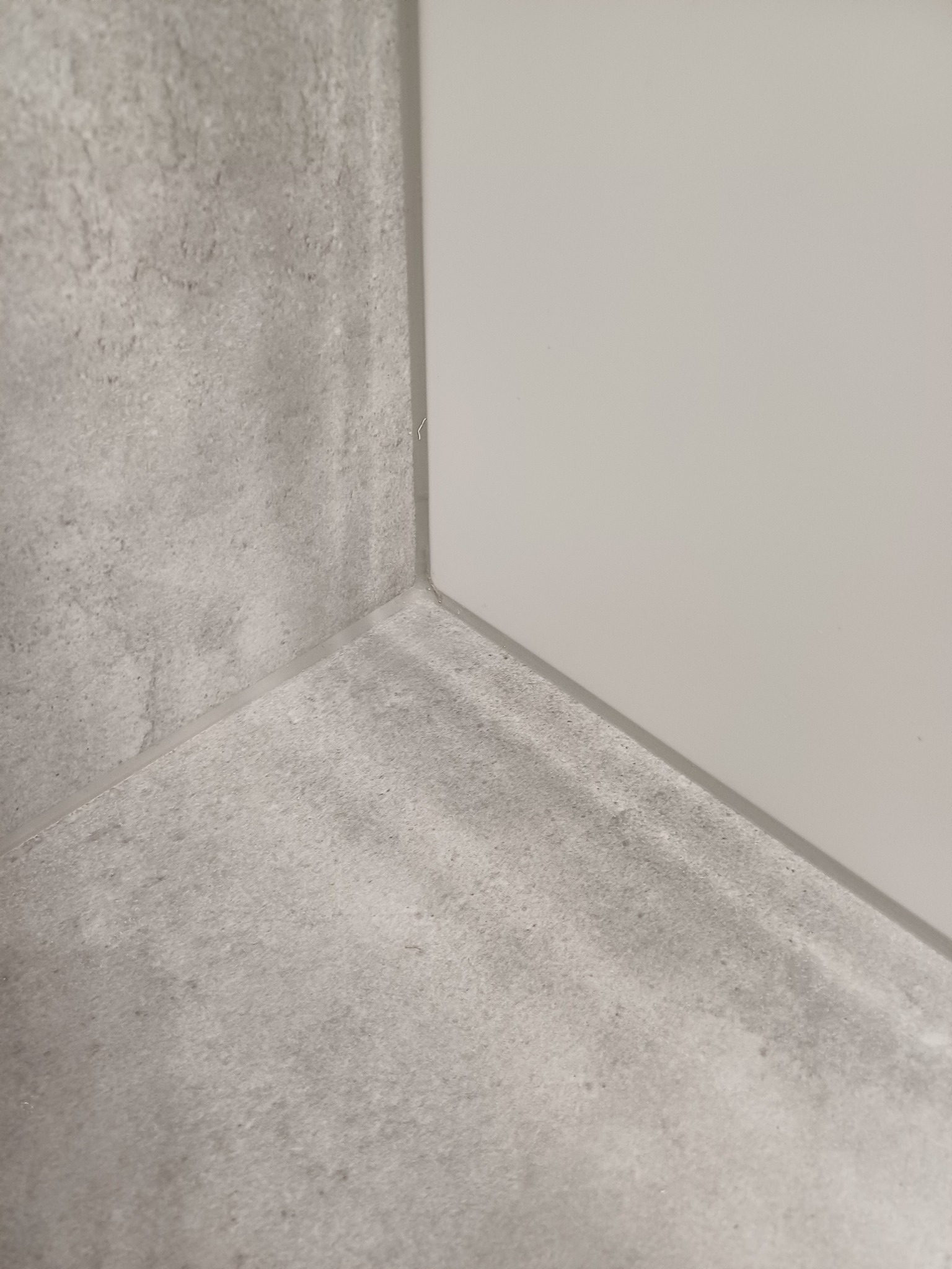 Szczegółowe ujęcie narożnika wykończonej ściany i podłogi w łazience, z widocznymi szarymi płytkami imitującymi beton i jasną, gładką powierzchnią, prawdopodobnie panelu lub płyty ściennej.