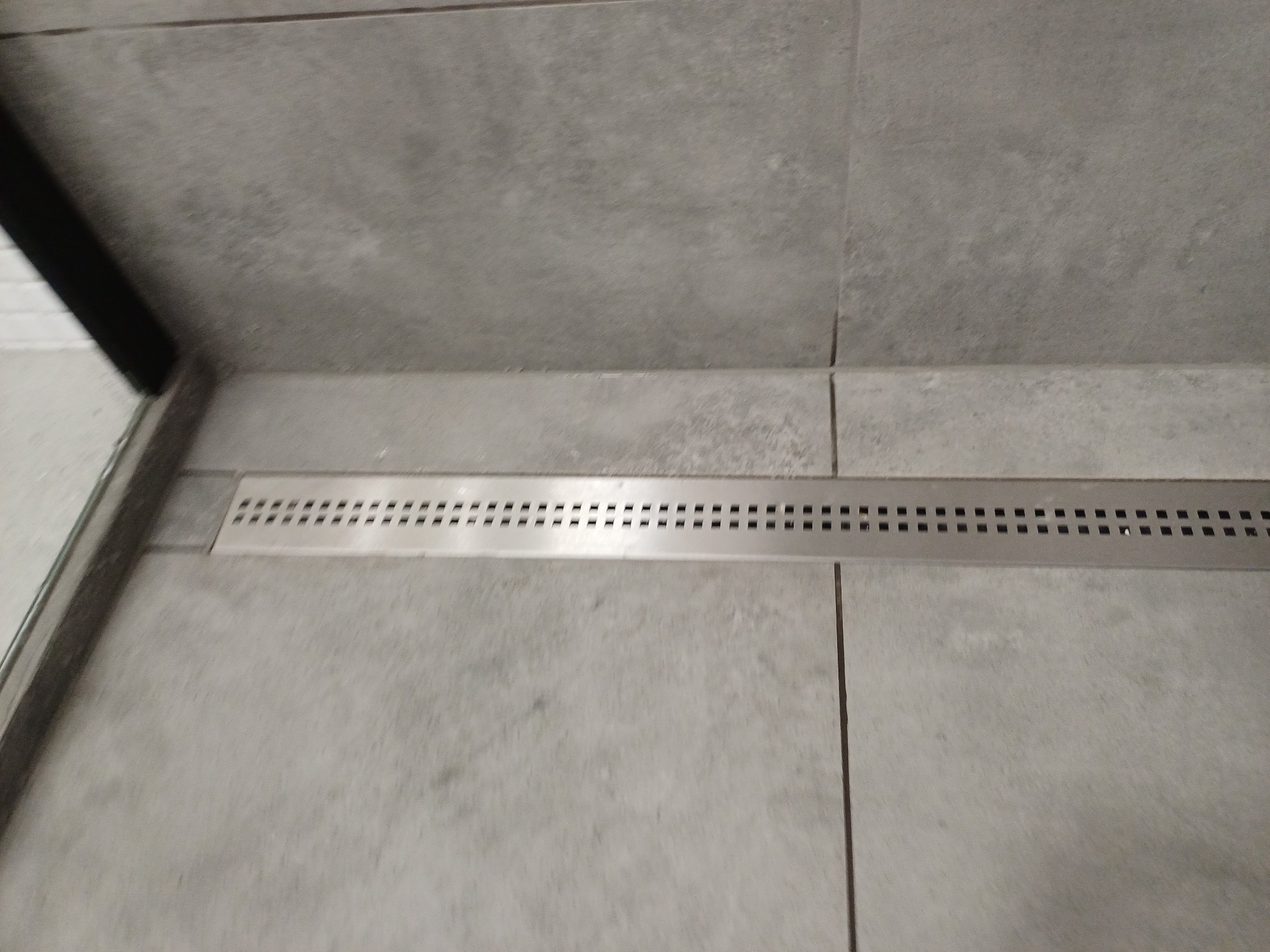 Szczegółowe ujęcie liniowego odpływu prysznicowego wkomponowanego w szare płytki podłogowe, z widocznym fragmentem kabiny prysznicowej po lewej stronie.