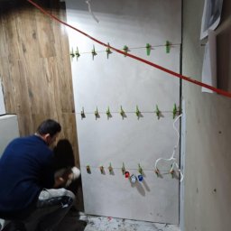 Karol Koksa - Układanie płytek imitujących drewno na ścianie w łazience, widoczne poziomice sznurkowe z klipsami, pracownik w trakcie nakładania kleju na płytkę.