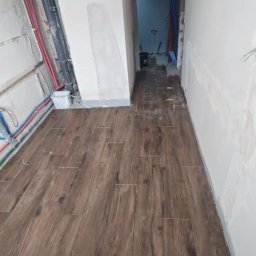 Karol Koksa - Układanie brązowych płytek podłogowych imitujących drewno w korytarzu podczas remontu mieszkania, widoczne wystające instalacje i ślady po szpachlowaniu na ścianach.