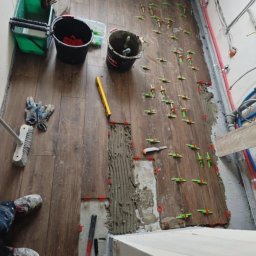 Karol Koksa - Układanie brązowych płytek podłogowych w łazience, widoczne narzędzia, poziomica, wiadro z klejem, krzyżyki dystansowe, fragment ściany po szpachlowaniu oraz osoba w roboczych ubraniach.