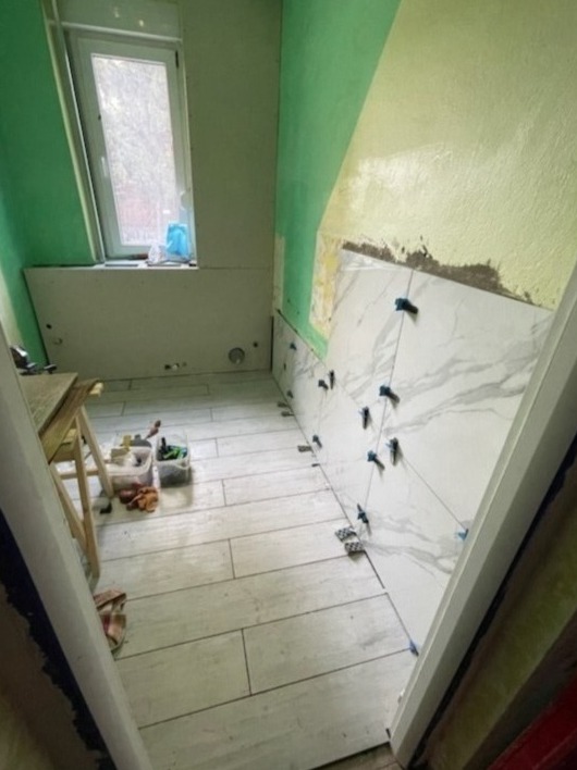 Wnętrze łazienki w trakcie układania płytek imitujących marmur na ścianie, widoczne krzyżyki dystansowe, podłoga wyłożona płytkami drewnopodobnymi, ściany częściowo pomalowane na zielono, przy...