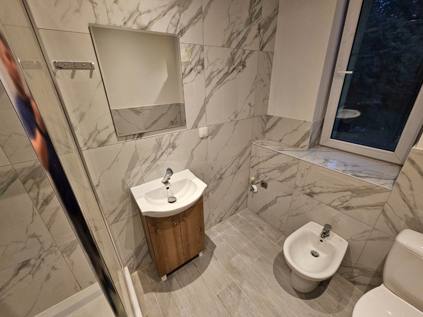 Wykończona łazienka w apartamencie z marmurowymi płytkami, drewnianą szafką pod umywalką i bidetem przy oknie.