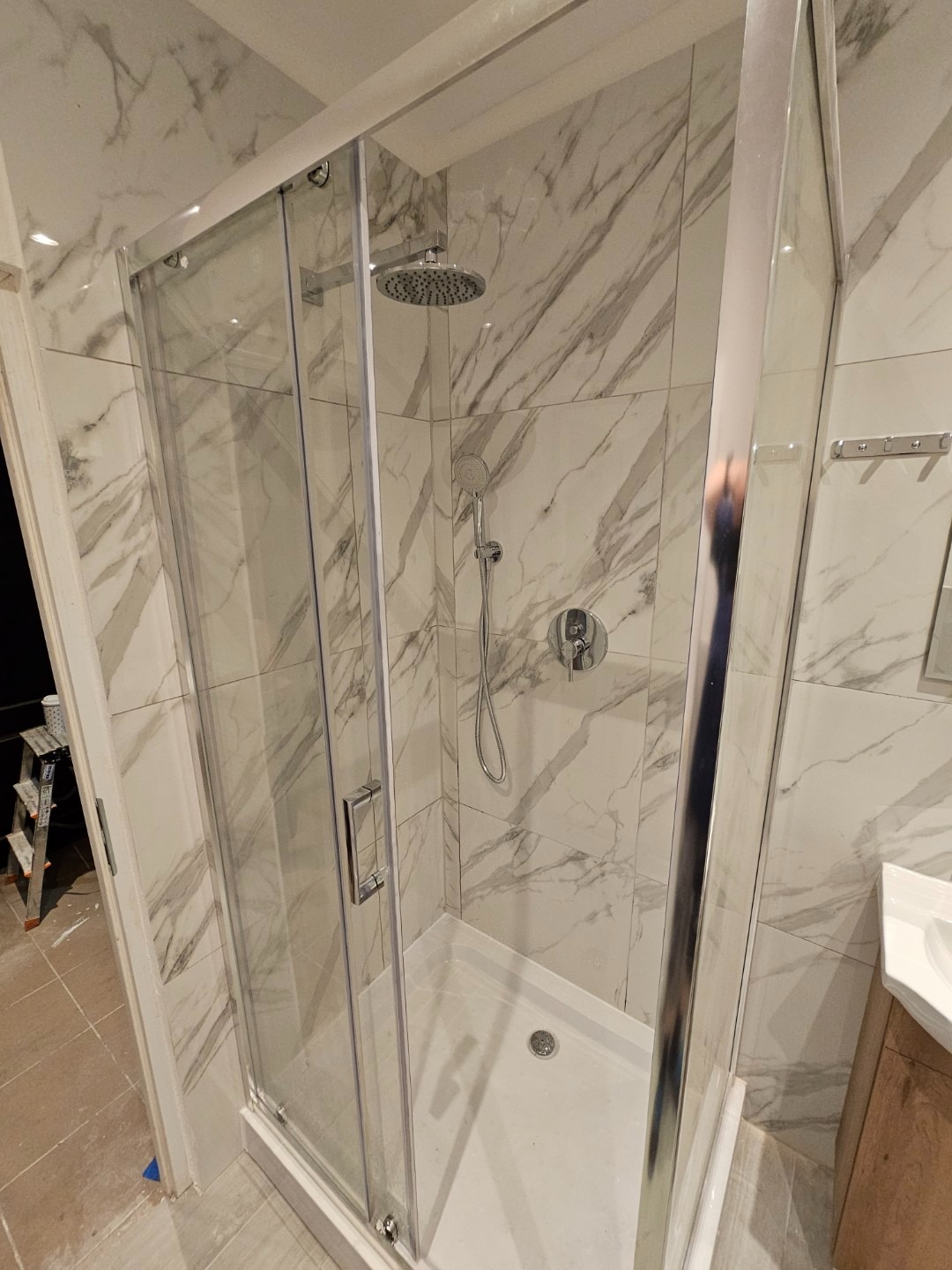 Nowoczesna kabina prysznicowa z przesuwnymi drzwiami, wykończona płytkami imitującymi marmur, z widoczną armaturą i odpływem liniowym; w tle widoczny fragment drabiny malarskiej.