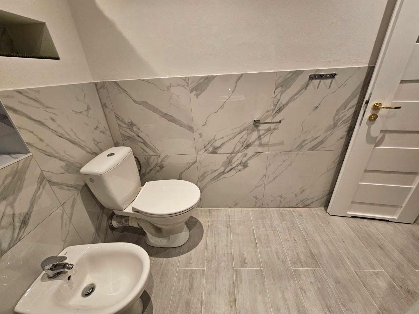 Nowoczesna łazienka w apartamencie z marmurowymi płytkami, białym sedesem i bidetem, drewnopodobna podłoga, widoczny fragment białych drzwi z złotą klamką.