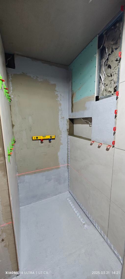 Wnętrze łazienki w trakcie prac: poziomica laserowa wyznacza linię, częściowo położone jasne płytki, zielone klipsy dystansowe, widoczne instalacje wodne, przygotowane ściany pod płytki.