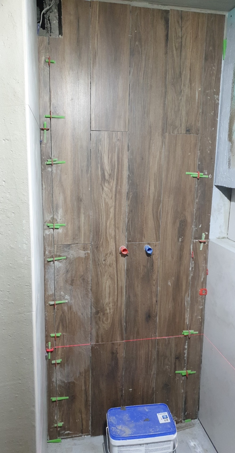 Ściana w trakcie układania płytek imitujących drewno, z widocznymi krzyżykami dystansowymi i poziomnicą laserową, niebieskie wiaderko z klejem na podłodze.