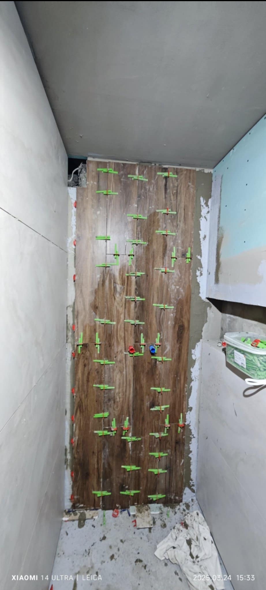 Montaż płytek imitujących drewno na ścianie w łazience, z użyciem zielonych krzyżyków dystansowych i czerwonych klinów poziomujących, widoczne ślady zaprawy na podłodze i ścianach.