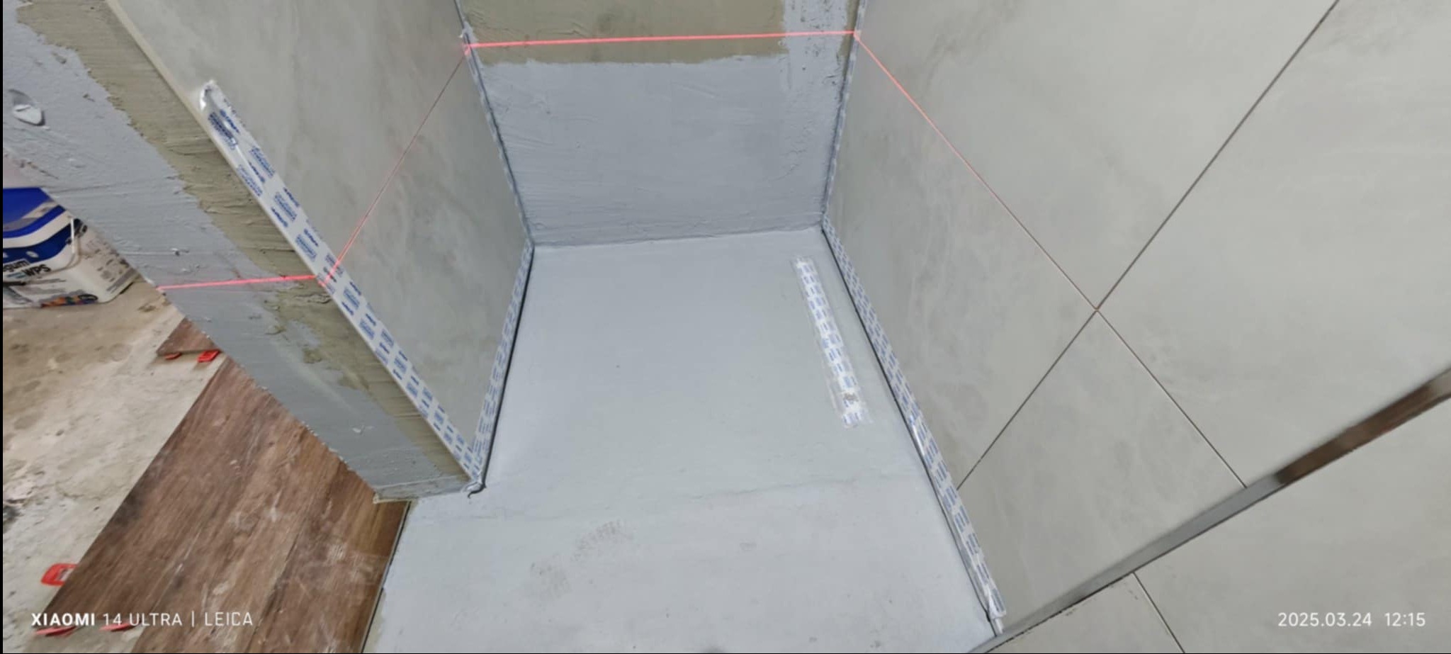 Przygotowanie do układania płytek w łazience, widoczne listwy startowe, poziomica laserowa wyznaczająca linię, surowy beton na ścianach.