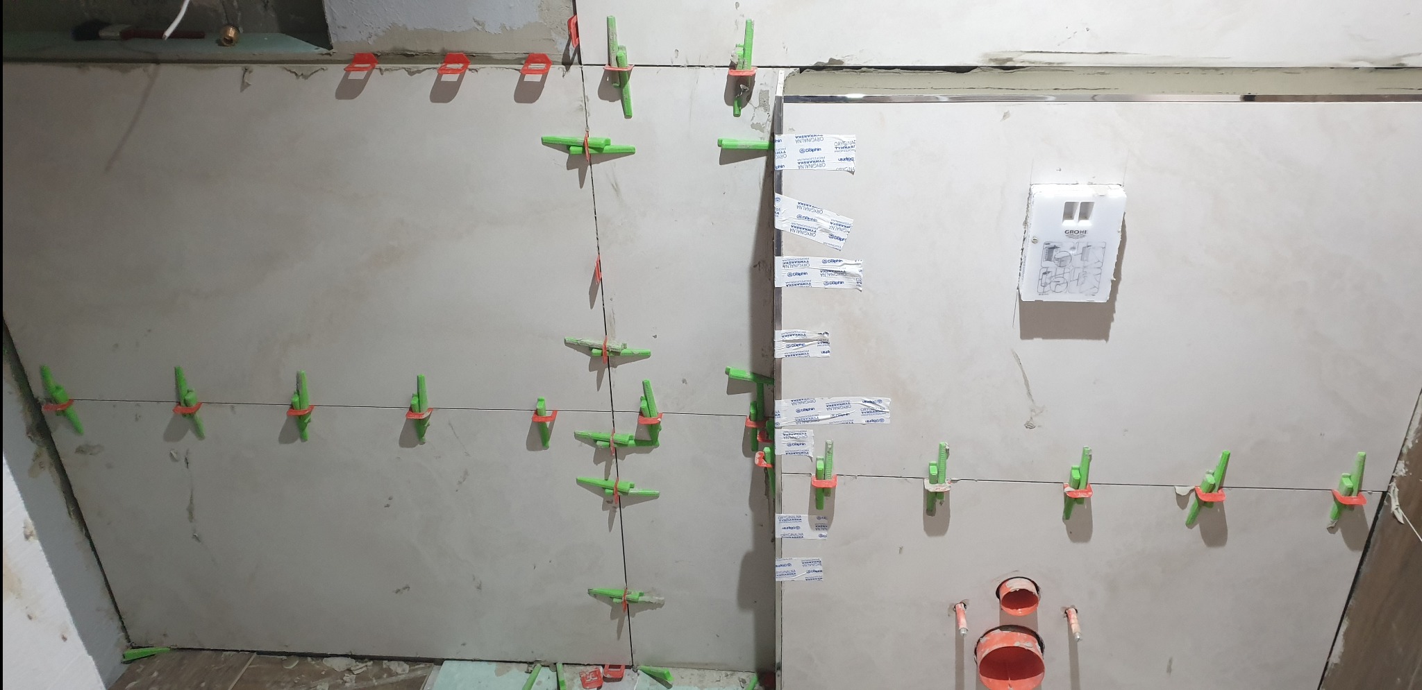 Układanie płytek w łazience z użyciem systemu poziomowania, widoczne otwory na instalacje wodne oraz schemat instalacji Grohe na ścianie.