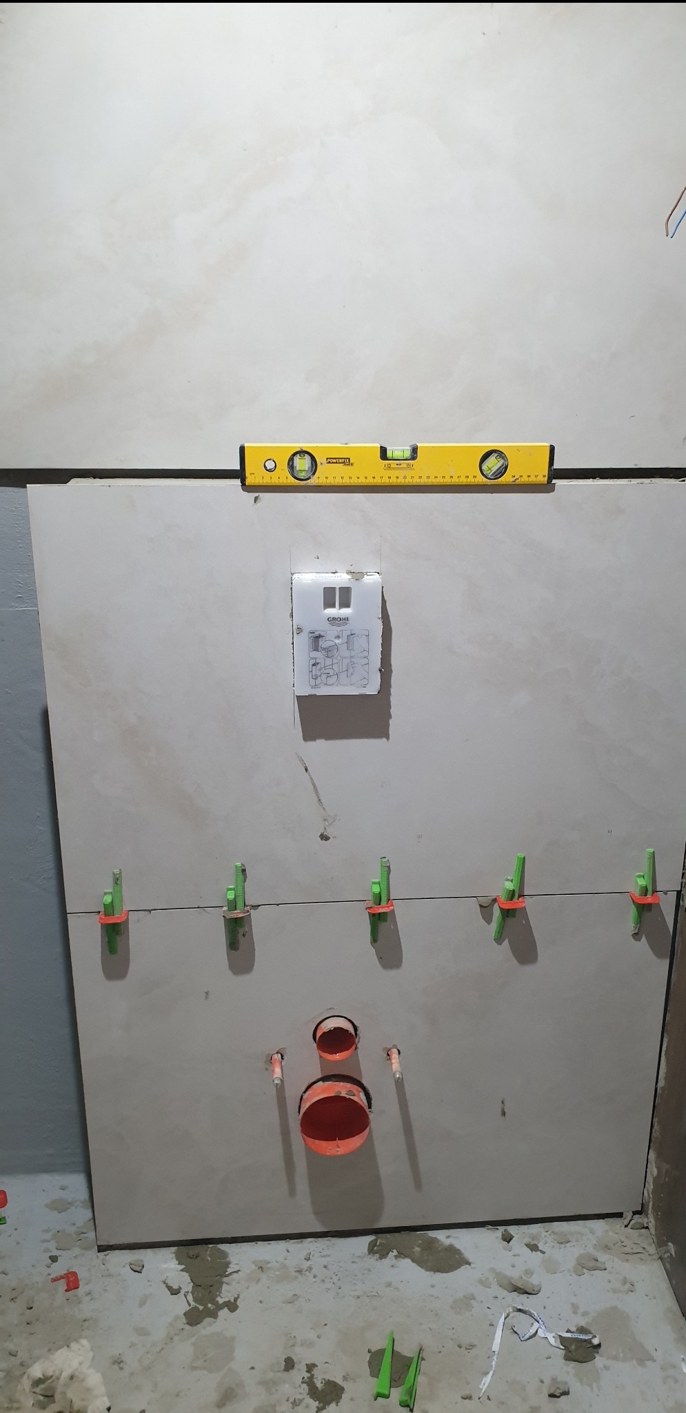 Montaż płytek ceramicznych w łazience, widoczny poziomica, system poziomowania płytek z klipsami i klinami, oraz instalacja sanitarna z rurami kanalizacyjnymi w ścianie.