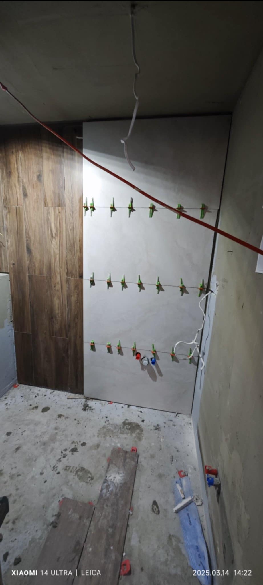 Układanie płytek imitujących drewno na ścianie w łazience, widoczne poziomice laserowe i krzyżyki dystansowe, surowy stan podłogi i wystające kable.