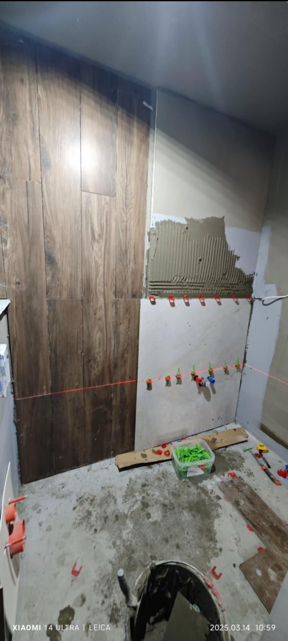 Układanie płytek imitujących drewno na ścianie w łazience, widoczne narzędzia, poziomica laserowa i pojemnik z klinami dystansowymi.