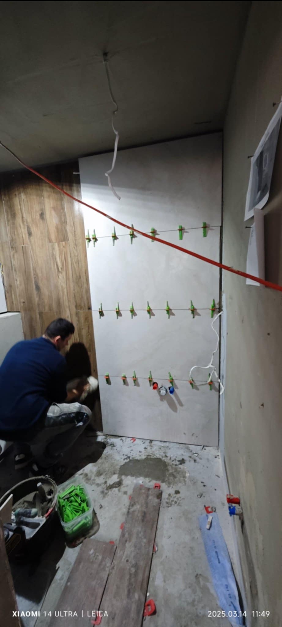 Układanie płytek imitujących drewno na ścianie w łazience, widoczne poziomice sznurkowe z klipsami, pracownik w trakcie nakładania kleju na płytkę.