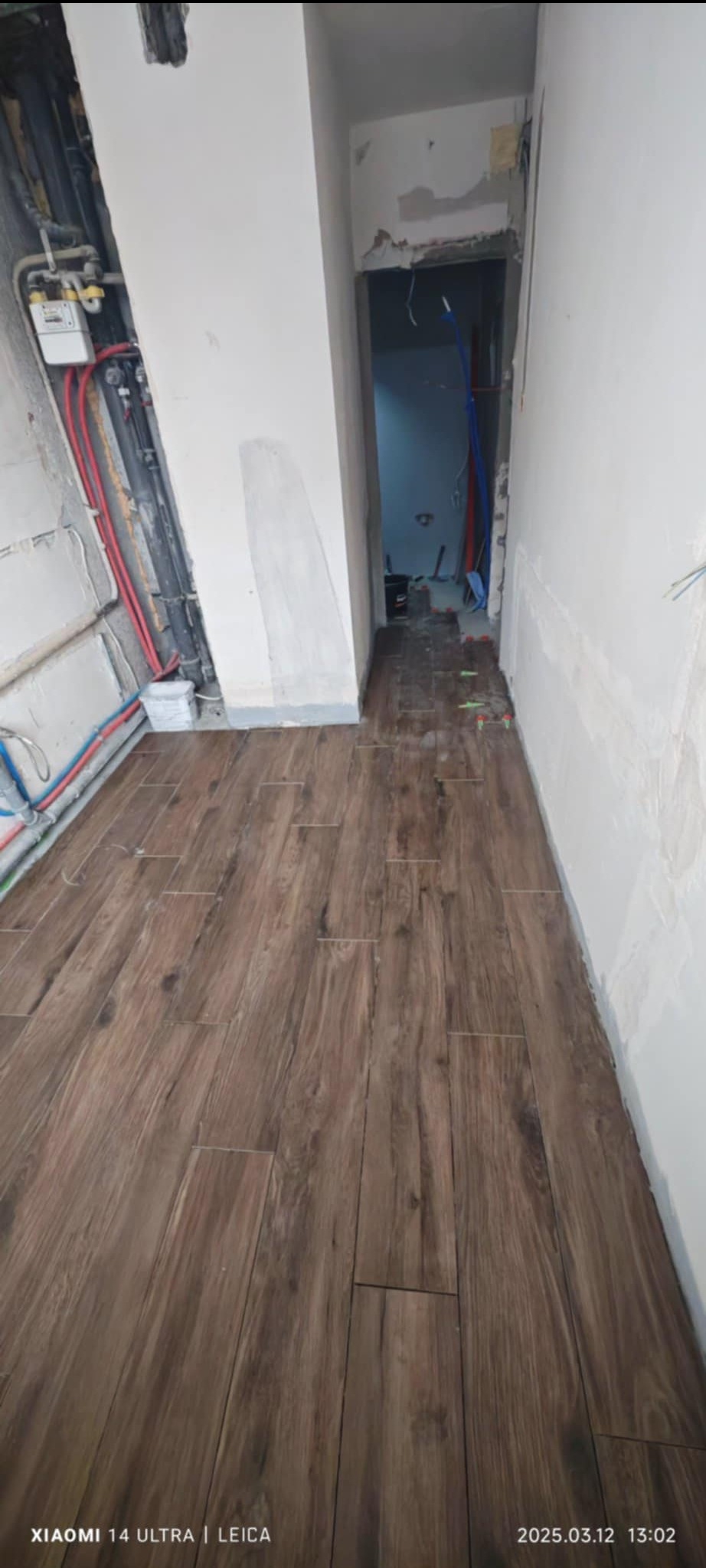 Układanie brązowych płytek podłogowych imitujących drewno w korytarzu podczas remontu mieszkania, widoczne wystające instalacje i ślady po szpachlowaniu na ścianach.