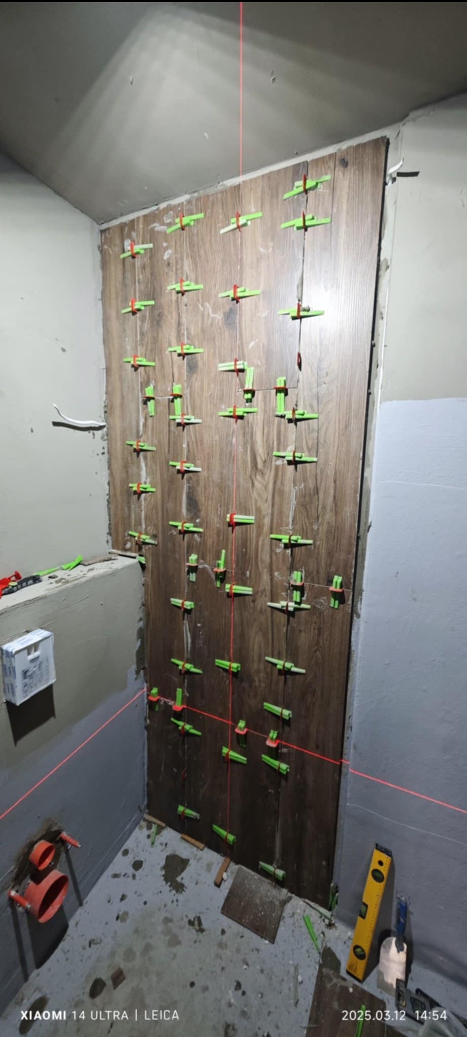 Układanie płytek imitujących drewno na ścianie w łazience, z użyciem poziomicy laserowej i klipsów poziomujących, widoczne fragmenty instalacji hydraulicznej.