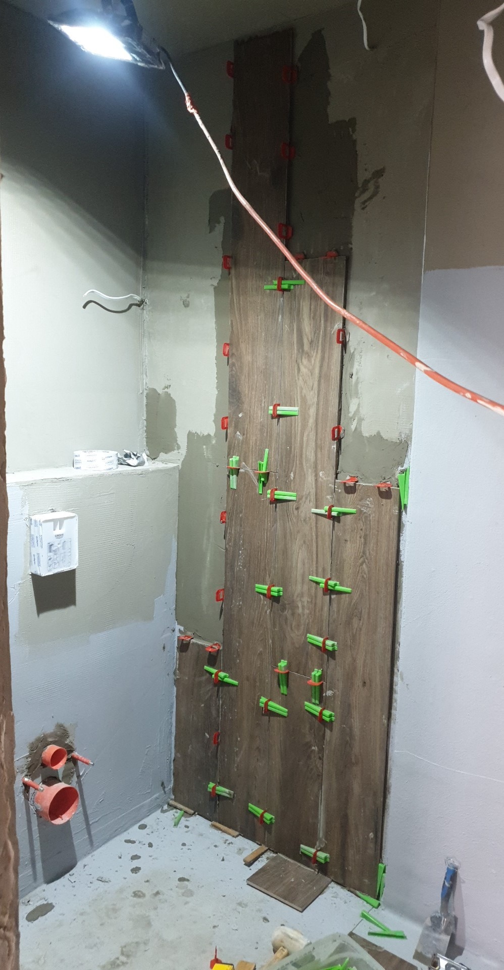Montaż płytek imitujących drewno na ścianie w łazience, widoczne krzyżyki dystansowe, przygotowane ściany i instalacja hydrauliczna.