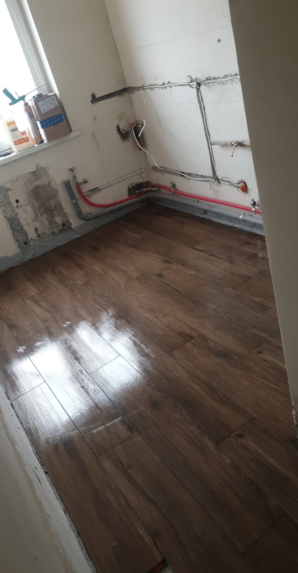 Nowo ułożona, błyszcząca podłoga z paneli drewnopodobnych w trakcie remontu łazienki, widoczne rury instalacji wodnej i przygotowane ściany do dalszych prac wykończeniowych, na parapecie okiennym...