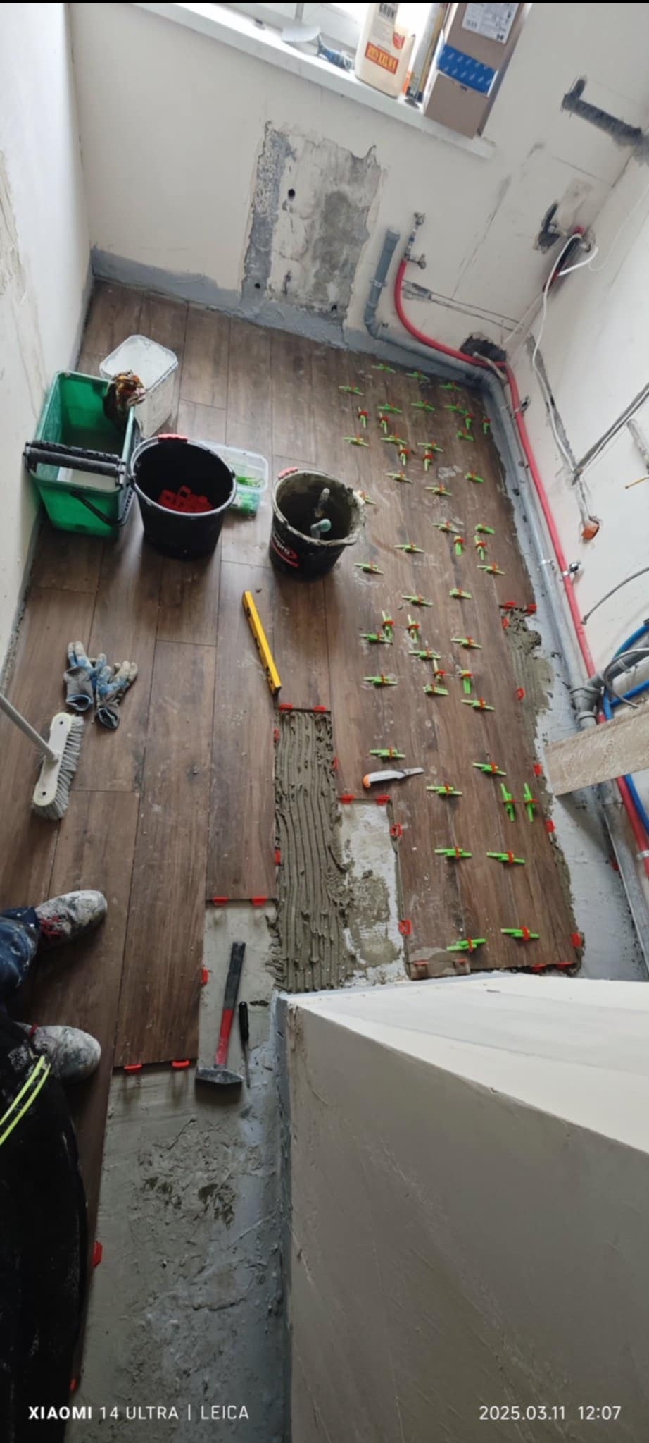 Układanie brązowych płytek podłogowych w łazience, widoczne narzędzia, poziomica, wiadro z klejem, krzyżyki dystansowe, fragment ściany po szpachlowaniu oraz osoba w roboczych ubraniach.