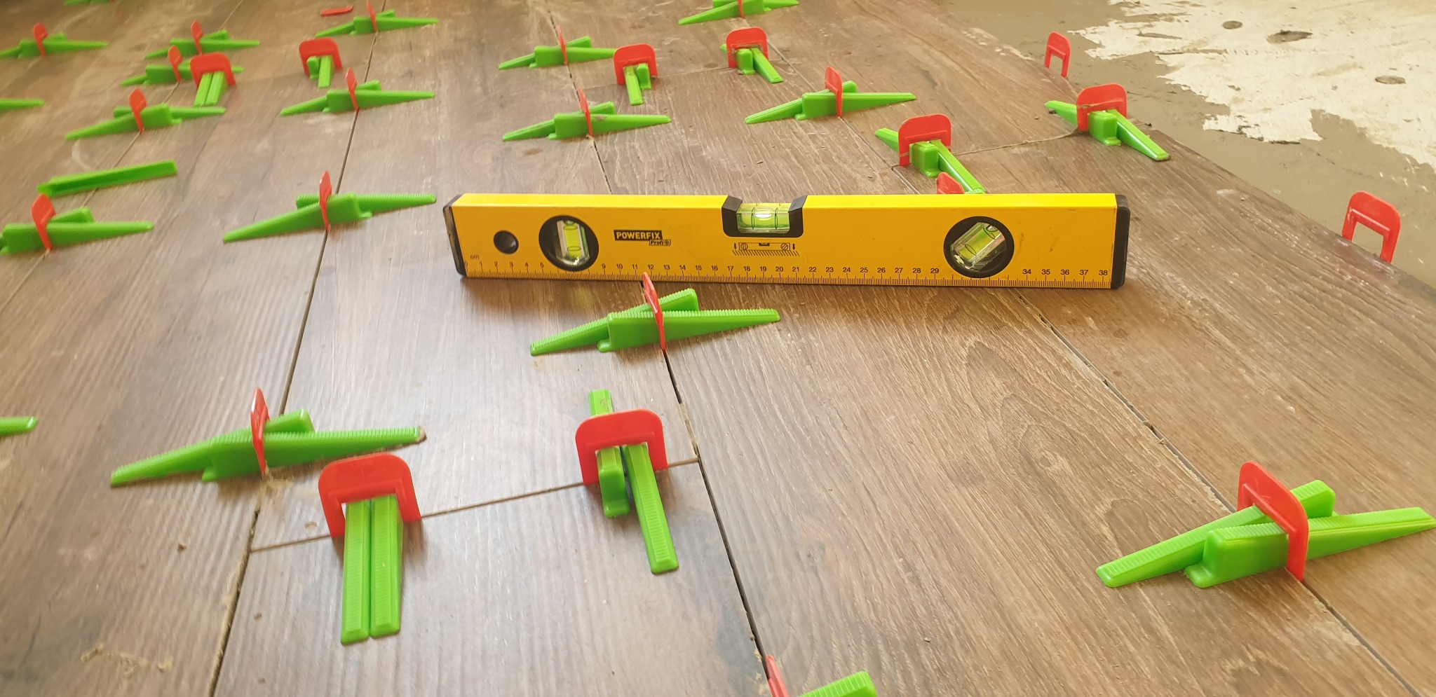 Układanie płytek podłogowych z użyciem poziomicy i systemu poziomowania płytek z zielonymi klinami i czerwonymi klipsami dystansowymi.