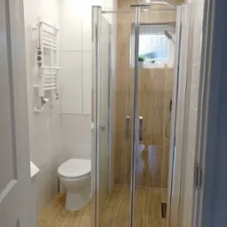 Nowoczesna łazienka z kabiną prysznicową z przezroczystego szkła, grzejnikiem drabinkowym i toaletą w neutralnych kolorach, wykończona płytkami imitującymi drewno na podłodze i części ściany.