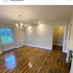 Pusta przestrzeń w jasnych kolorach z odnowioną podłogą z ciemnego drewna, widok na okno z zielenią na zewnątrz i ozdobny żyrandol.