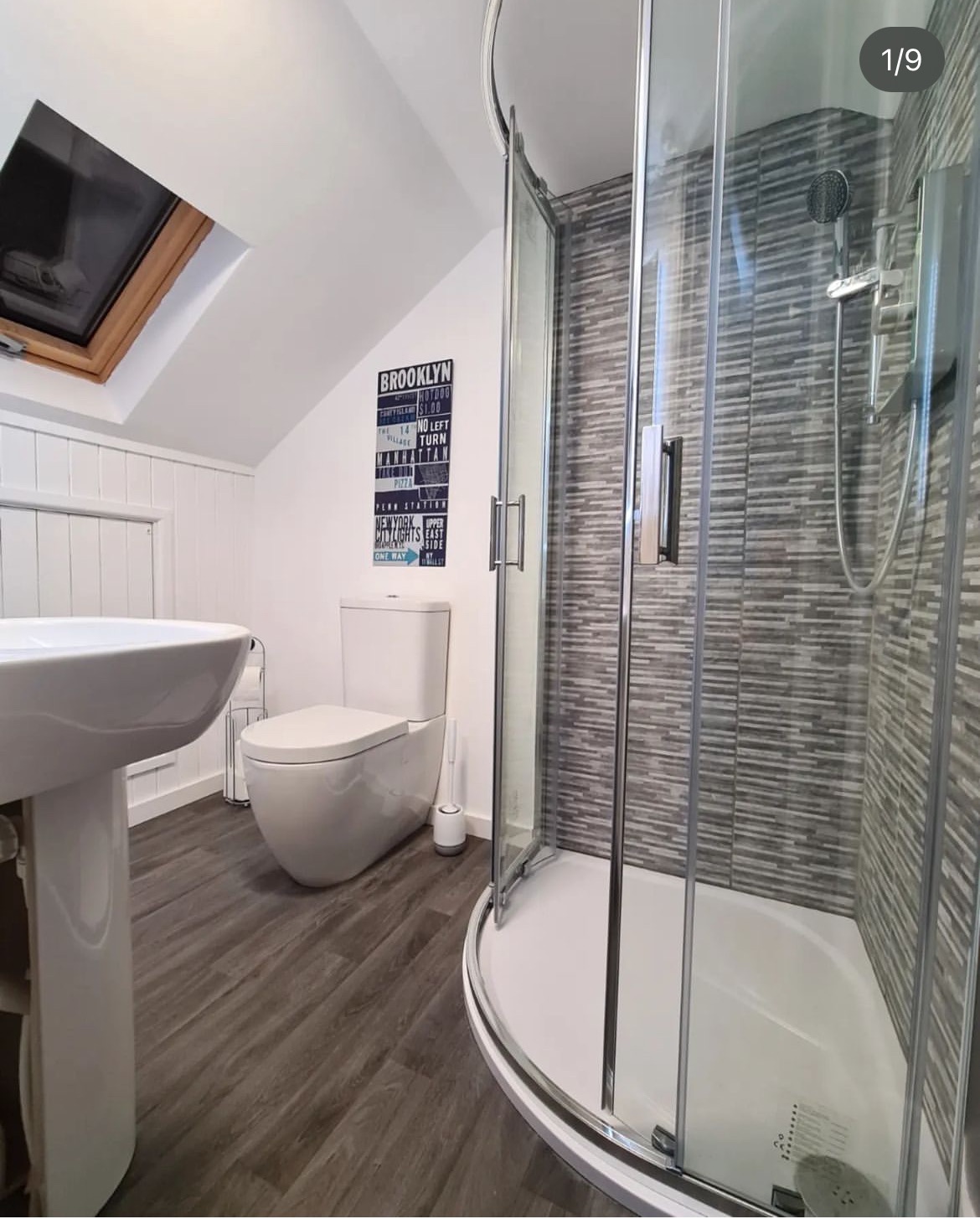 Nowoczesna łazienka na poddaszu z kabiną prysznicową półokrągłą, umywalką na postumencie, toaletą i ciemną podłogą imitującą drewno. Na ścianie plakat w stylu vintage z motywami nowojorskimi.