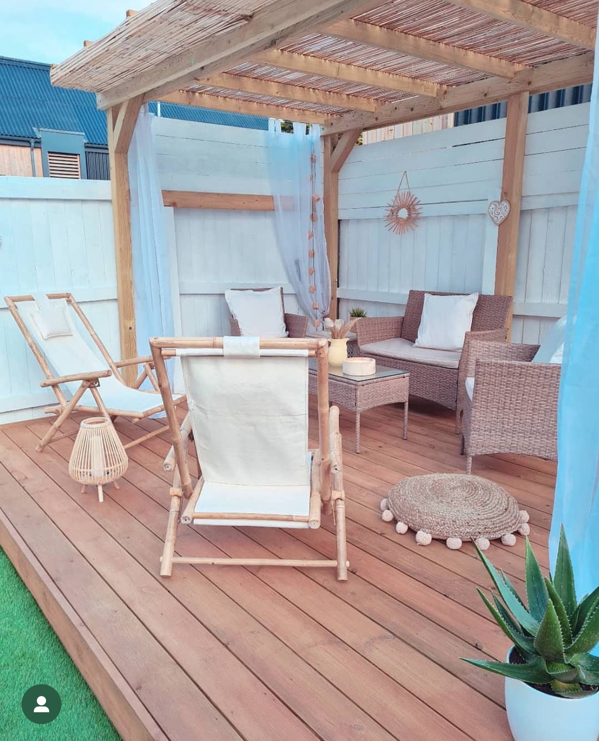 Drewniany taras z pergolą, rattanowymi meblami ogrodowymi i leżakami z jasnego płótna, udekorowany zwiewnymi, białymi zasłonami i roślinami w doniczkach.