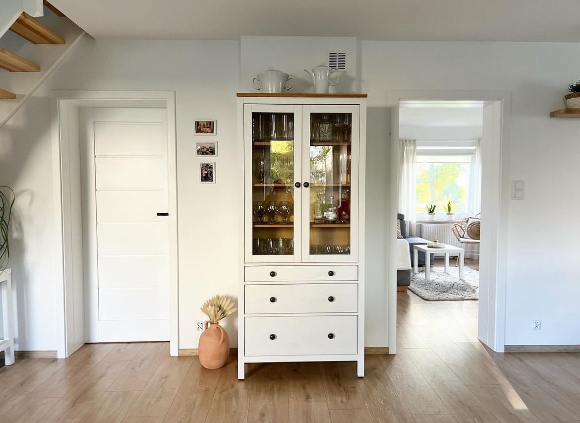 Jasne, przestronne wnętrze z drewnianą podłogą, białymi ścianami i białym kredensem z przeszkloną górną częścią, na którym stoją białe naczynia, a obok drzwi wisi galeria zdjęć, widok na salon...