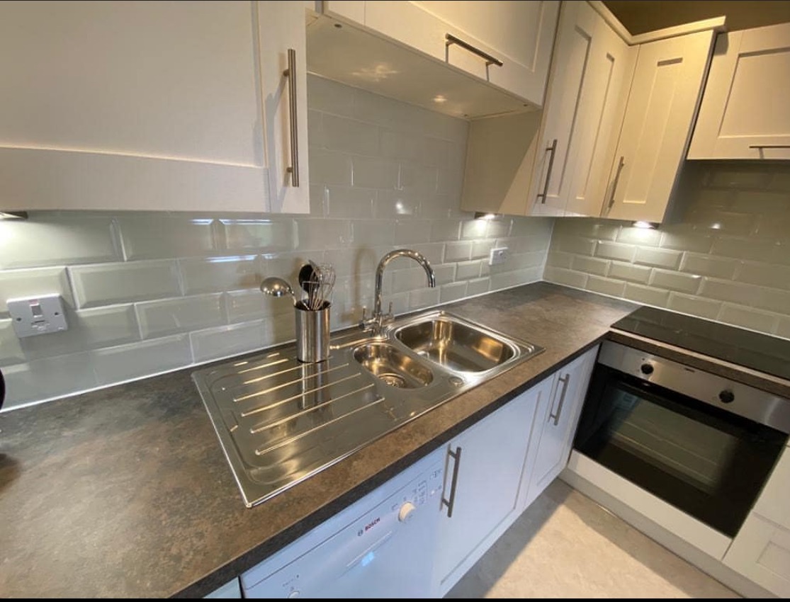 Nowoczesna kuchnia z białymi szafkami, szarym blatem i błyszczącymi, zielonkawymi płytkami na ścianie, widoczny zlewozmywak ze stali nierdzewnej, bateria i akcesoria kuchenne.