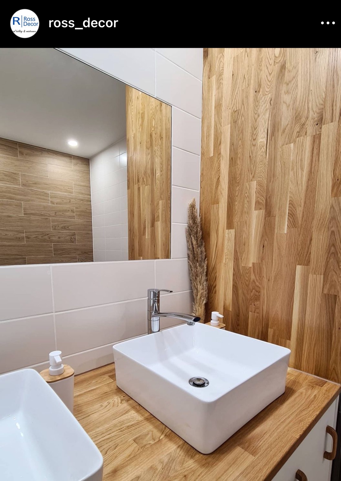 Nowoczesna łazienka z drewnianą ścianą, białą umywalką nablatową i dużym lustrem, odbijającym wnętrze z brązowymi płytkami imitującymi drewno.