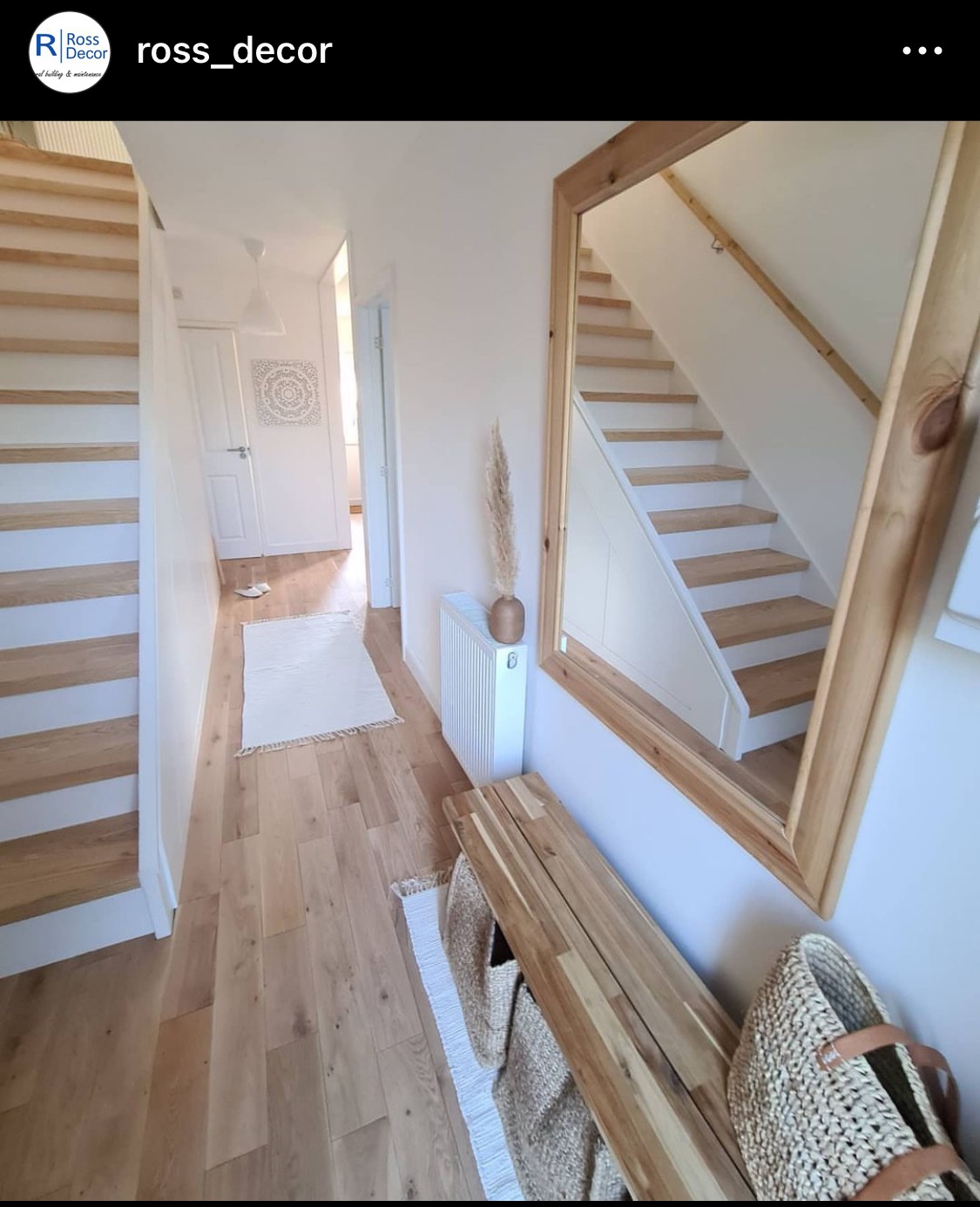 Jasny przedpokój w Ostaszewie z drewnianą podłogą, schodami z białymi podstopnicami, dużym lustrem w drewnianej ramie i drewnianą ławką z koszami.