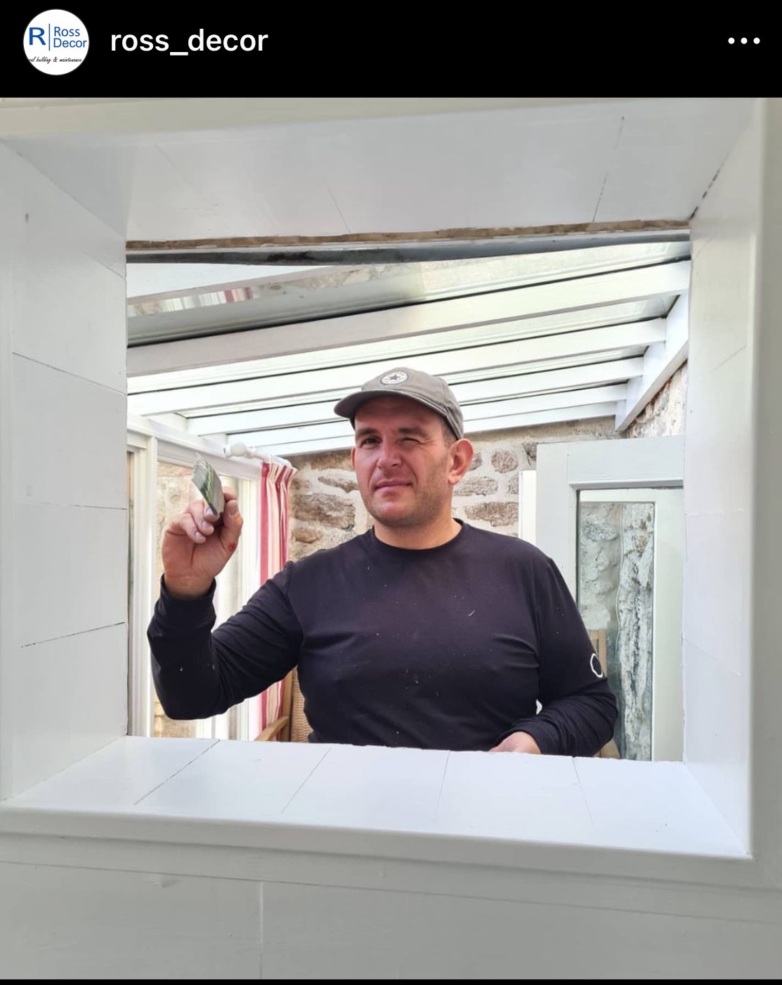 Mężczyzna w czarnej koszulce i czapce z daszkiem, mrugający okiem, trzymający pędzel malarski w przeszklonym pomieszczeniu z kamienną ścianą w tle, maluje na biało framugę okna.