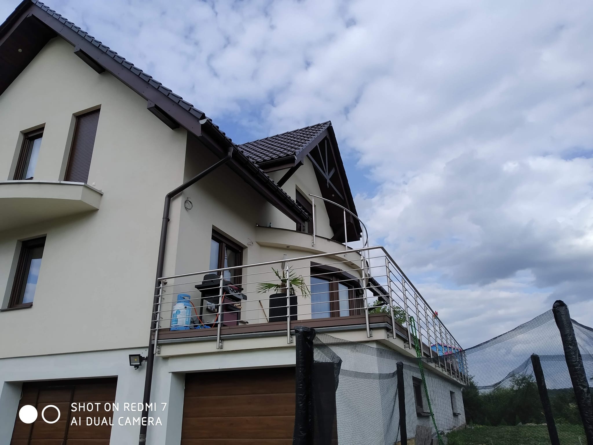 Dom z beżową elewacją i balkonem z balustradą ze stali nierdzewnej. Na balkonie grill i roślina w donicy. Widok z dołu, pochmurne niebo w tle.