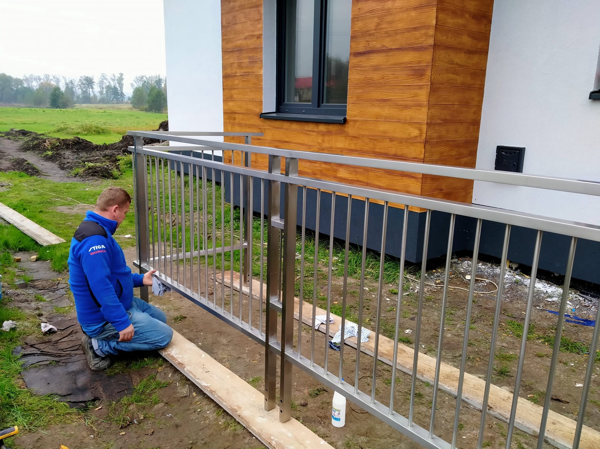 Montaż balustrady metalowej przy budynku z drewnianą elewacją. Pracownik czyści konstrukcję, widoczne narzędzia i elementy montażowe. Tło stanowi zielony trawnik.